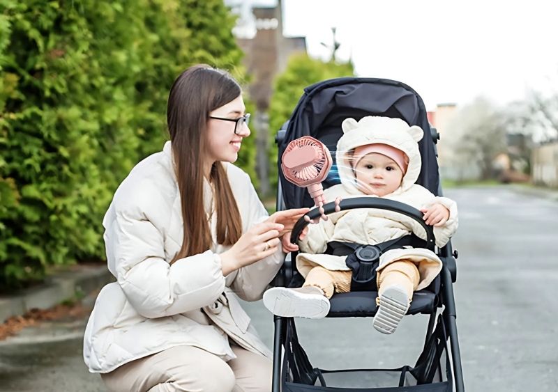 Một bé gái dùng quạt mini gắn vào xe đẩy khi đi dạo công viên cùng cha mẹ